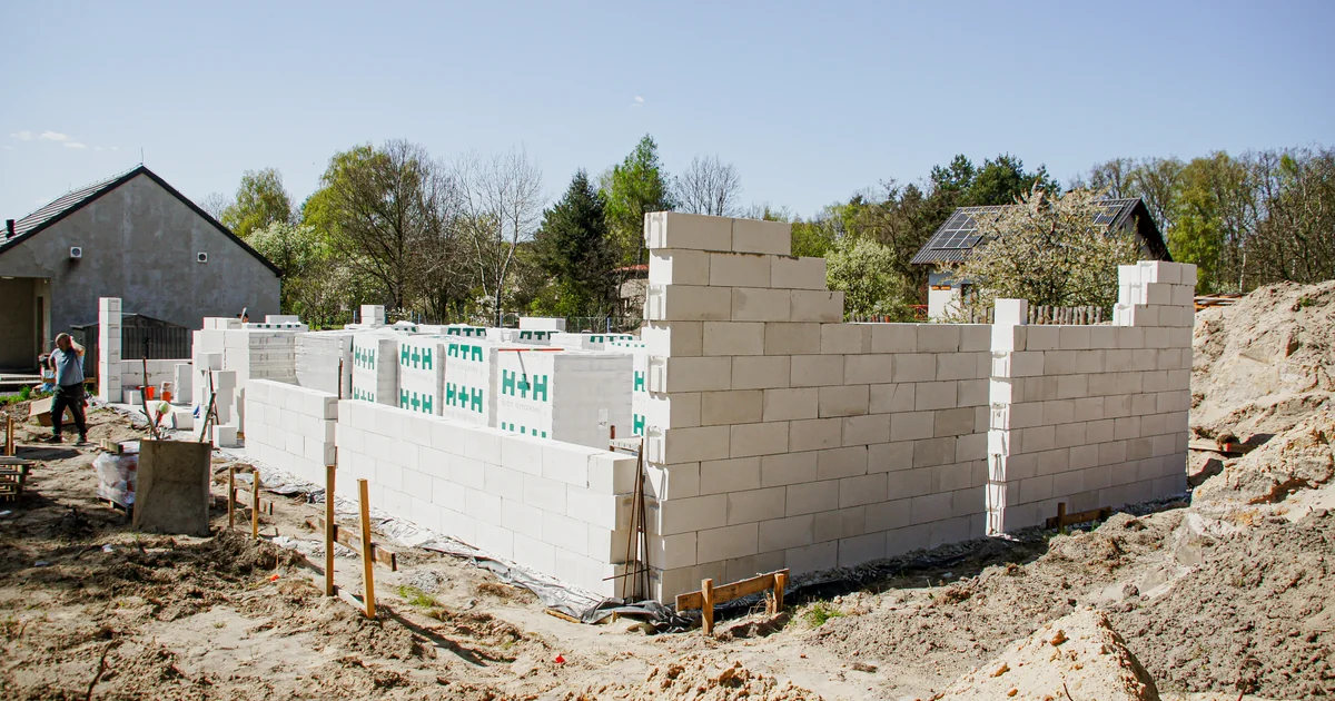Budowa domu jednorodzinnego CoreLTB Builders w Mikołowie-Zgody (kwiecień 2026) - mury z betonu komórkowego H+H w trakcie murowania, ciepła płyta fundamentowa 30 cm, zielone palety H+H z materiałem, IV kategoria górnicza. Realny etap SSO widziany od zewnątrz, wiosenne słońce, kontekst osiedla mieszkaniowego.